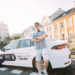 Młody mężczyzna w okularach przeciwsłonecznych stoi przy białym samochodzie Uber Comfort. Chcesz wiedzieć, jak zostać kierowcą Ubera?